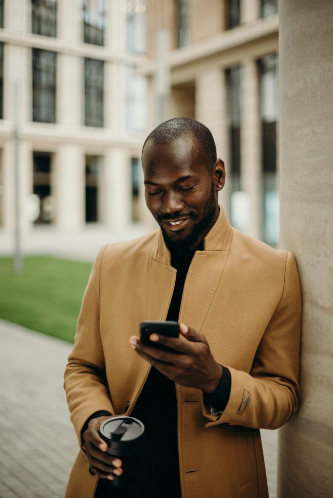 Relaxed man in coat smiles while using smartphone and holding coffee outside. Social Media.