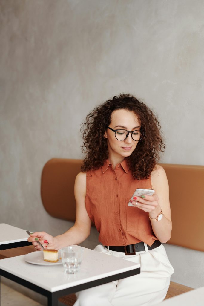 Curly-haired woman in a café checking her phone, enjoying cheesecake.